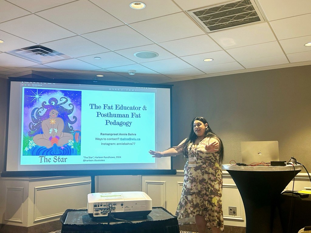 image of me presenting at a conference. I am standing on the right wearing a print wrap dress and lanyard. My arms are holding out as I talk with my hands. My hair is falling down my shoulders/back. To the left of me is a projector screen that has a slide sharing a image of the tarot "star" card created by @harleen.illustrates and title of my presentation "The Fat Educator & Posthuman Fat Pedagogy". At the bottom of the image is a projector.